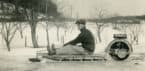 Young man sits on a motorized sled steered with a tiller; trees, fence posts, and power lines in the background.